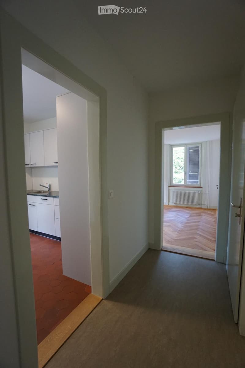 Empty hallway with light-colored walls and wooden flooring, leading to a kitchen and another room with wooden flooring and a window.