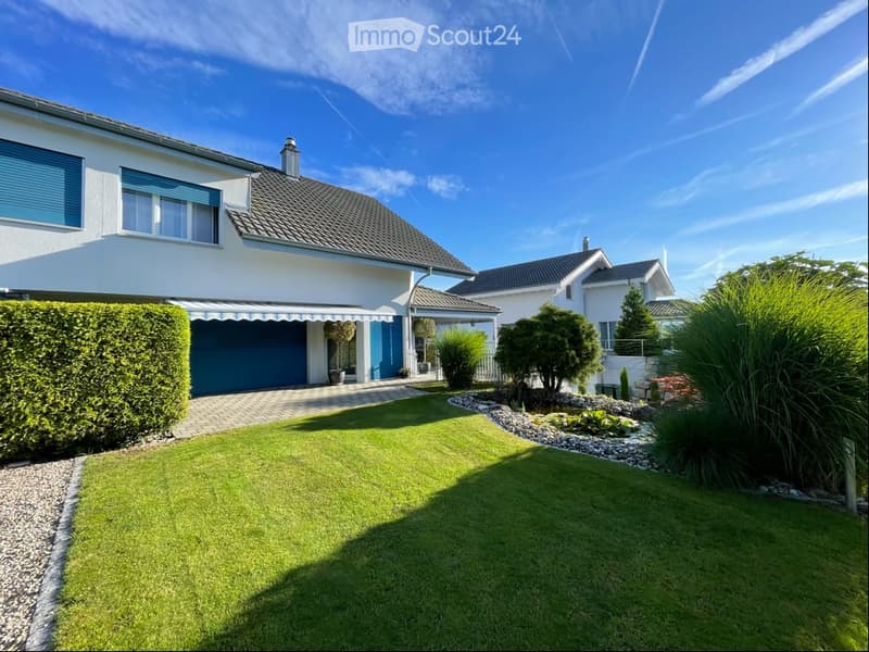 A modern two-story house with white walls, blue shutters, and a grey tiled roof. The house features a well-maintained lawn, a small garden area with shrubs and plants, and a driveway leading to a garage.