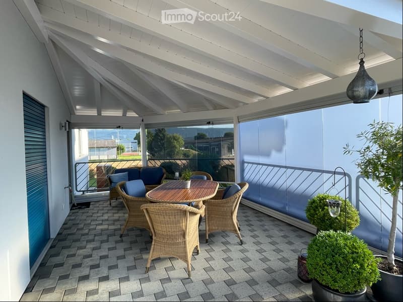 A modern, covered balcony with a wooden table, wicker chairs, and potted plants, overlooking a scenic view.