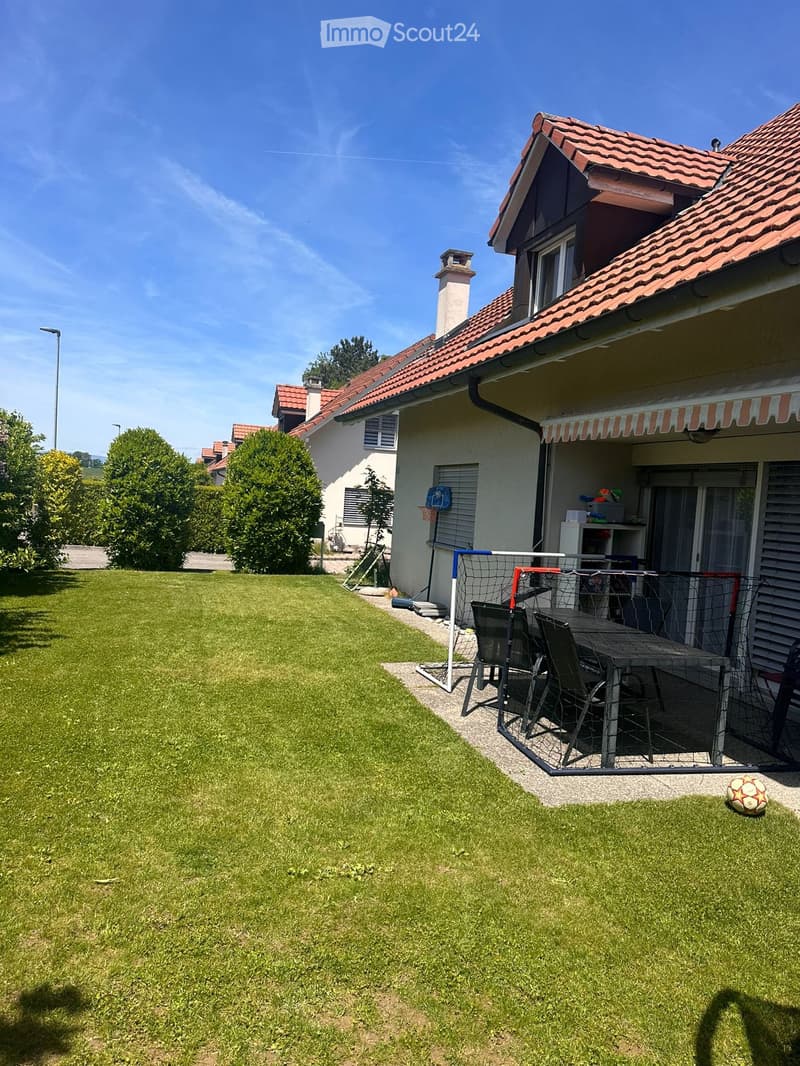Well-maintained lawn with a patio area featuring a table, chairs, a small goal, and a soccer ball. The house has a red-tiled roof and is surrounded by trees.