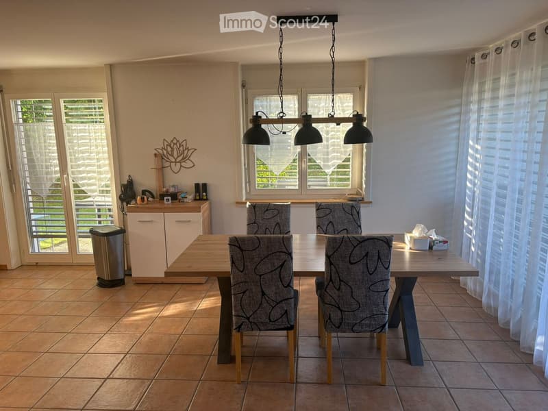 Dining room with a wooden dining table, four patterned chairs, a white cabinet, and a trash can. The room has tiled flooring, large windows with curtains, and a hanging light fixture.