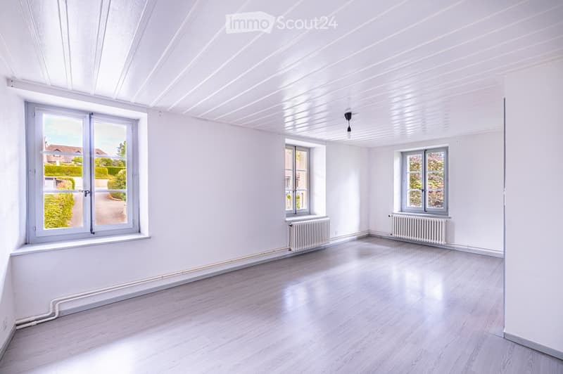 Empty room with white walls, light wooden flooring, and large windows allowing natural light. Radiators are present under the windows, and the ceiling has a modern, ribbed design.