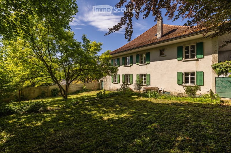 A two-story house with a red-tiled roof and green shutters, surrounded by a lush garden with trees and a bench.