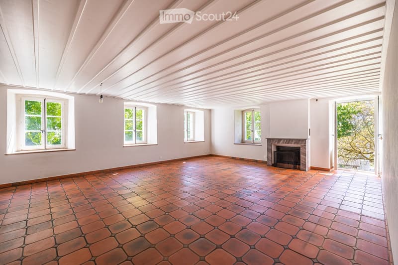 Empty room with exposed wooden ceiling beams, terracotta tile floor, multiple windows, and a fireplace with a black insert.