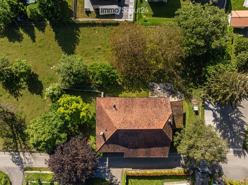 Aerial view of a two-story house with a brown tiled roof, surrounded by a well-maintained lawn and trees. The house has multiple chimneys and is situated near a road with neighboring houses visible in the background.