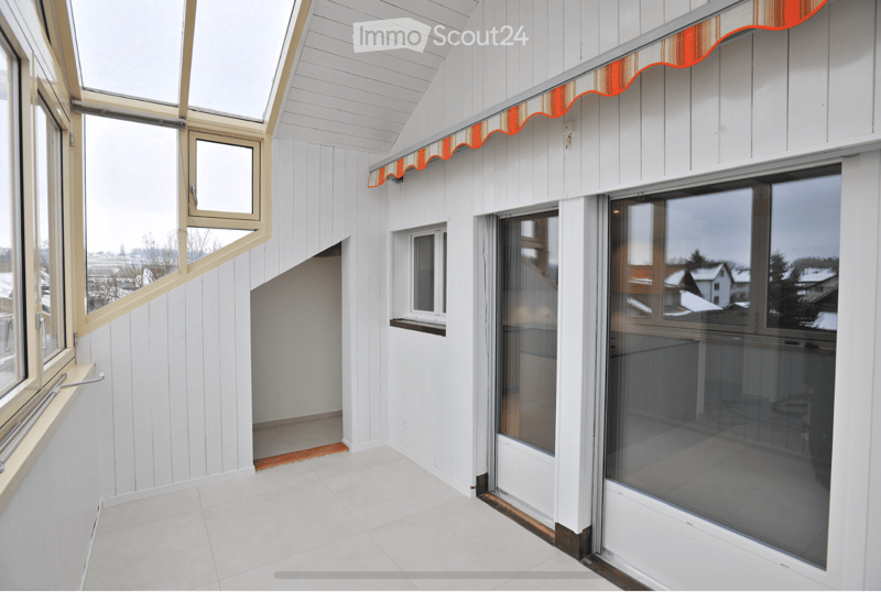 Bright, modern balcony with white wooden paneling, large glass windows, and a red and white striped awning. The space is clean and minimalistic with a tiled floor.