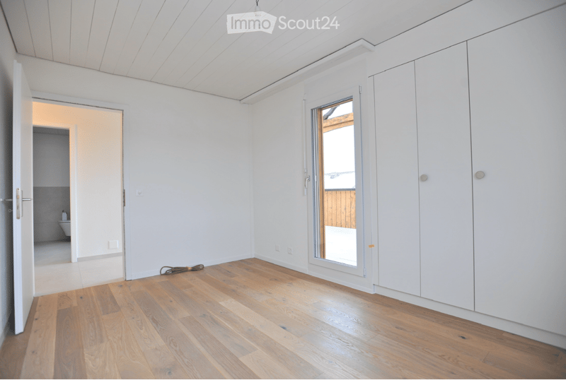 Empty room with white walls, wooden flooring, built-in white cabinets, and a large window with a view of an outdoor area. A small pile of cables is on the floor.
