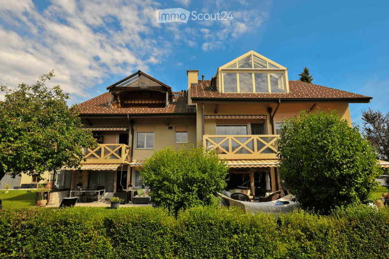 Two-story house with a tiled roof, large windows, and a balcony. The house features a well-maintained garden with outdoor furniture and greenery.