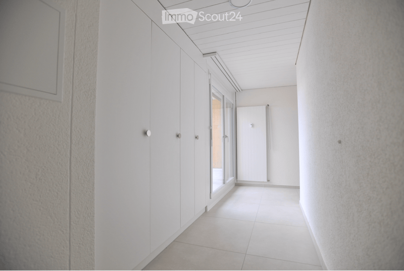 A modern, minimalist hallway with white walls, white tiled flooring, and built-in white cabinetry on one side. A large glass door at the end leads to another space.