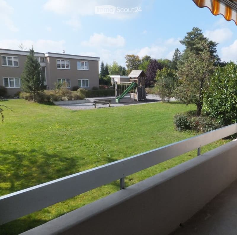 Balcony overlooking a green lawn with a playground, a bench, and a building in the background.