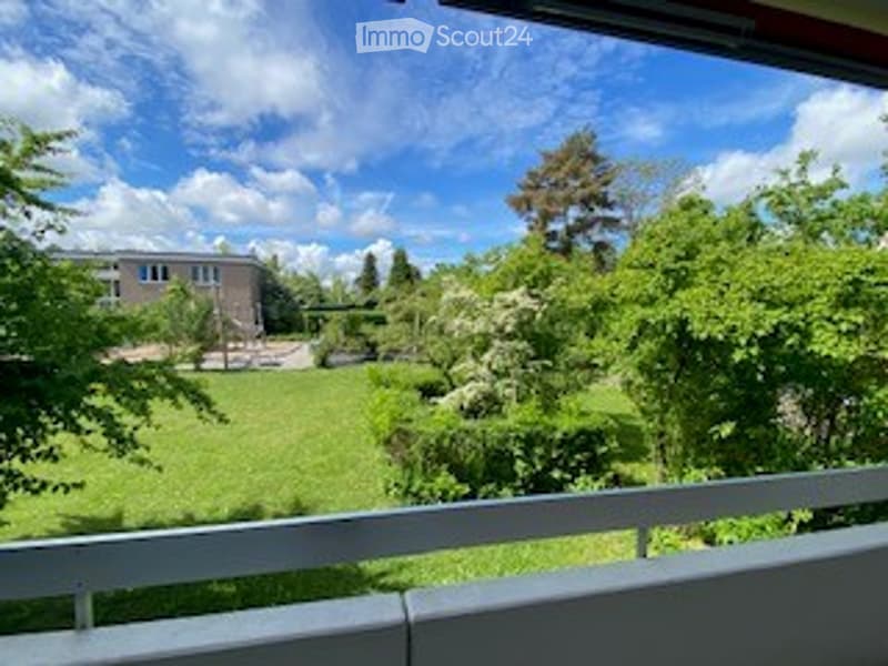 Balcony overlooking a green lawn and trees, with a building visible in the distance under a partly cloudy sky.