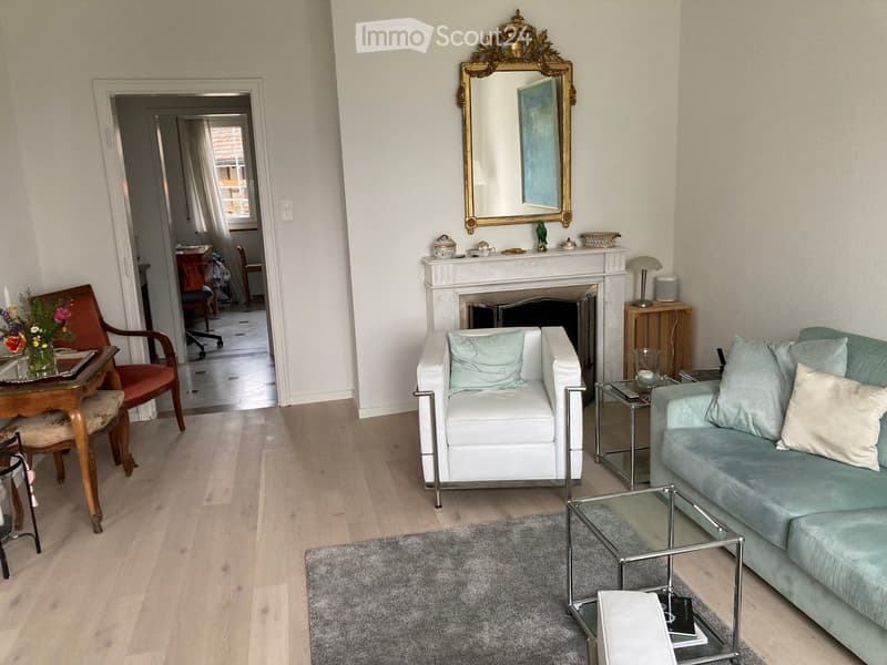 Elegant living room with light-colored hardwood flooring, a white sofa, a light blue armchair, a glass coffee table, a fireplace with decorative items, and a large ornate mirror above it.
