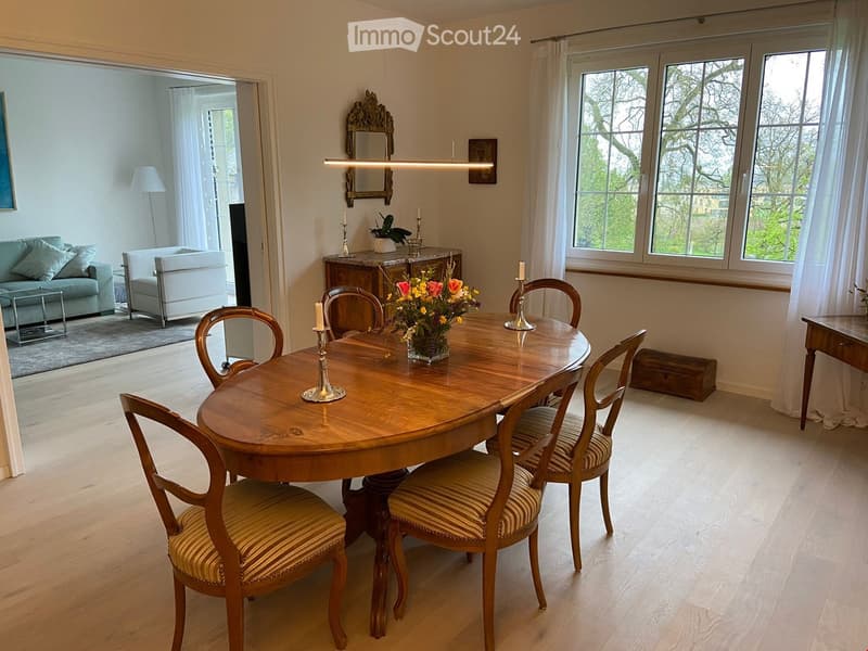 Elegant dining room with a round wooden table, six chairs, a flower vase, candles, a sofa, a table lamp, and a mirror on the wall.