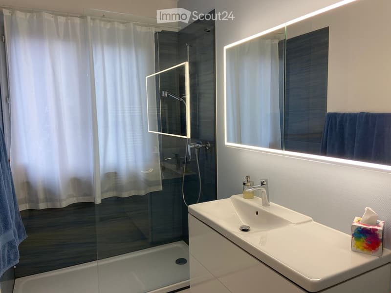 Modern bathroom with a white sink, chrome faucet, large illuminated mirror, glass shower stall with white curtain, and blue towel.