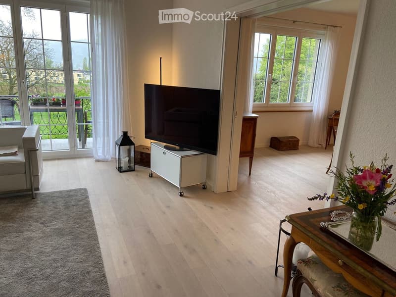 Bright living room with large windows and a view, featuring a grey sofa, flat-screen TV on a stand, wooden coffee table with a flower vase, and a small side table with a chair.