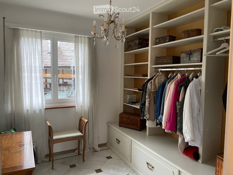 A well-lit room with a large built-in wardrobe, shelves, a chandelier, a chair, and a window with curtains.