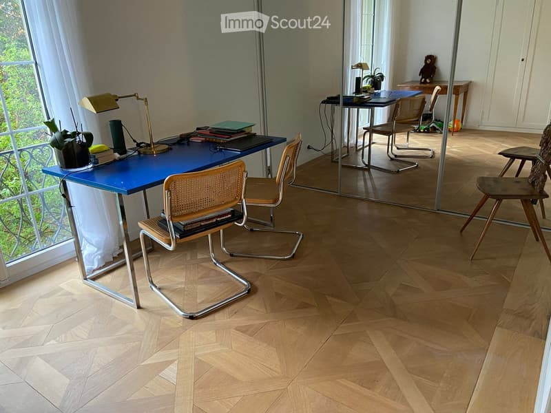 Modern office space with herringbone wooden flooring, large window with sheer curtains, blue desk, wicker chairs, and various office supplies.