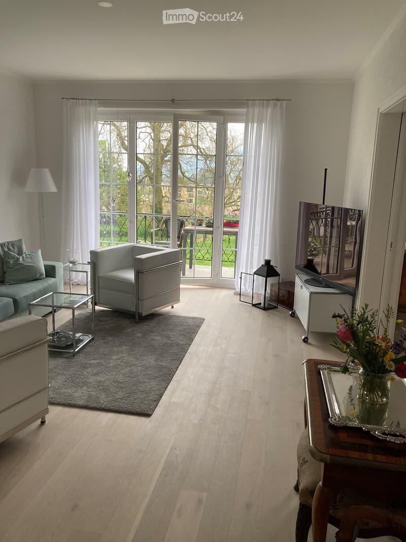 Modern living room with light wooden flooring, large windows with white curtains, a grey sofa, armchair, coffee table, and a TV stand with a mirror. A small table with a flower vase is near the TV stand, and a lantern is placed on the floor.
