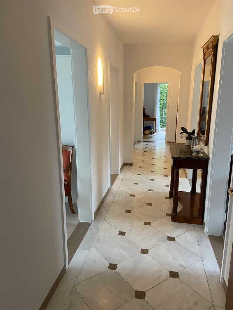 Elegant hallway with hexagonal tile flooring, white walls, and warm lighting. Features a small table with a vase and a mirror on the right side, and a glimpse of a room with a table and chairs at the end.