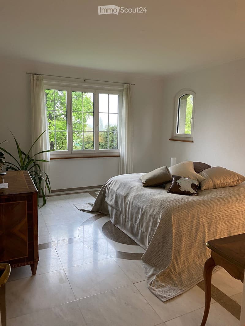 Bright bedroom with a large bed, multiple pillows, a nightstand, a chair, and a plant near the window. The room features large windows with curtains and a small round mirror on the wall.