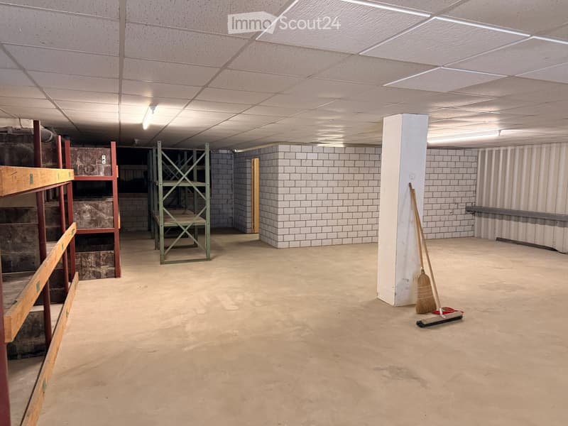 Empty basement room with concrete floor, white tiled walls, exposed ceiling lights, and a broom leaning against a pillar.