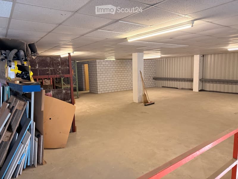 Empty room with concrete floor, white tiled walls, exposed ceiling with fluorescent lighting, and a broom leaning against a pillar. There are some stacked materials and a large cardboard box on the left side.