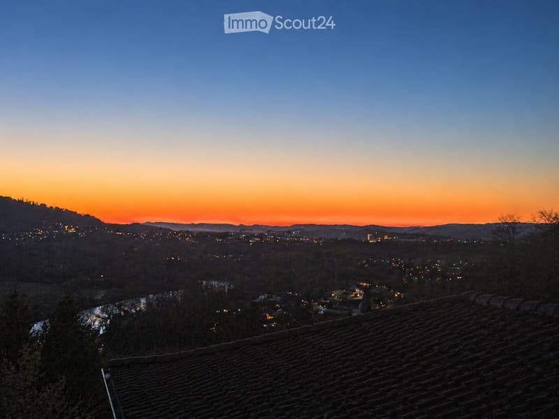 Sunset view from a terrace with a tiled roof, overlooking a town with lights and a river.