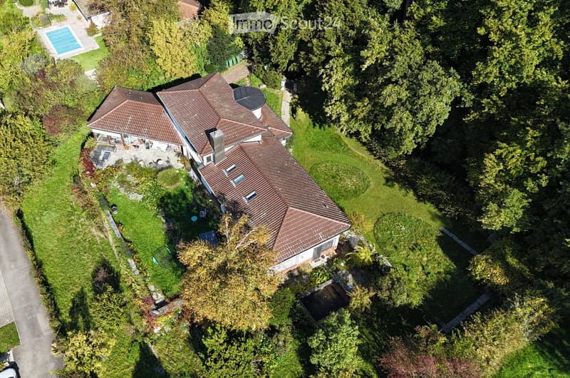 Aerial view of a house with a brown roof, surrounded by trees and grass. A swimming pool is visible in the distance.