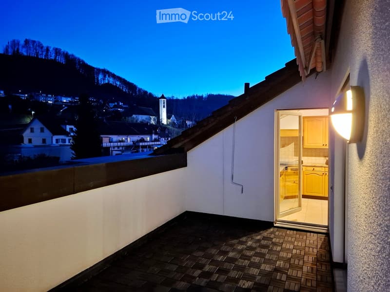 Balcony with tiled floor, open sliding door leading to a brightly lit kitchen with yellow cabinets, overlooking a town with visible houses and a church tower.