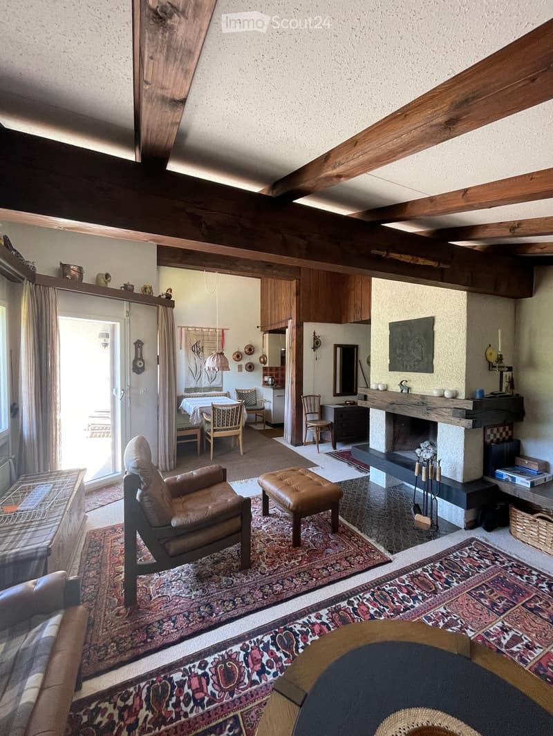 Cozy living room with exposed wooden beams, leather sofa, ottoman, coffee table, fireplace, and dining area visible through an open doorway.