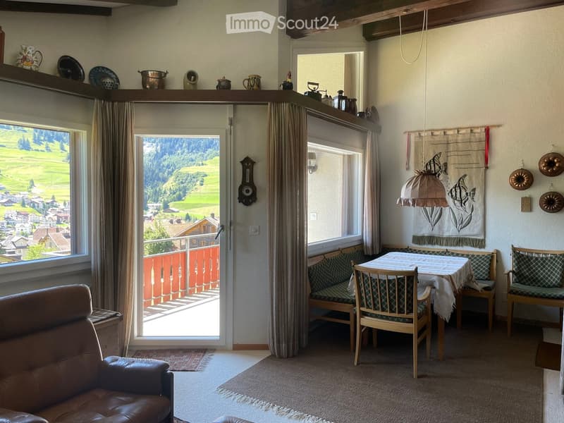Cozy living room with a leather sofa, dining table with chairs, bench seating, large windows with curtains, and a scenic mountain view through the open door to a balcony.