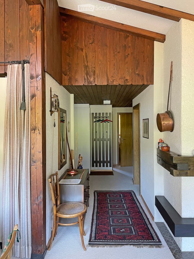 Cozy hallway with wooden beams, a patterned rug, a small wooden chair, and a small table with a bowl on it. Wooden doors and built-in wooden cabinets are visible.