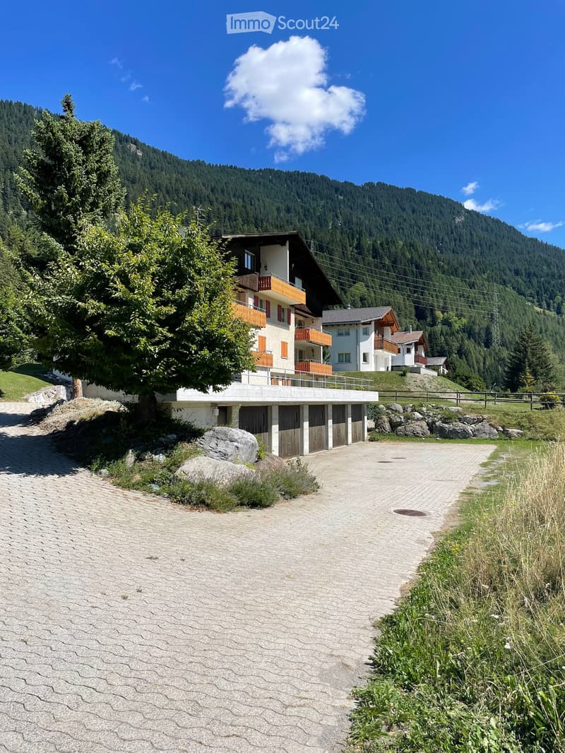 A two-story house with balconies, surrounded by trees and mountains, with a paved driveway and garage.