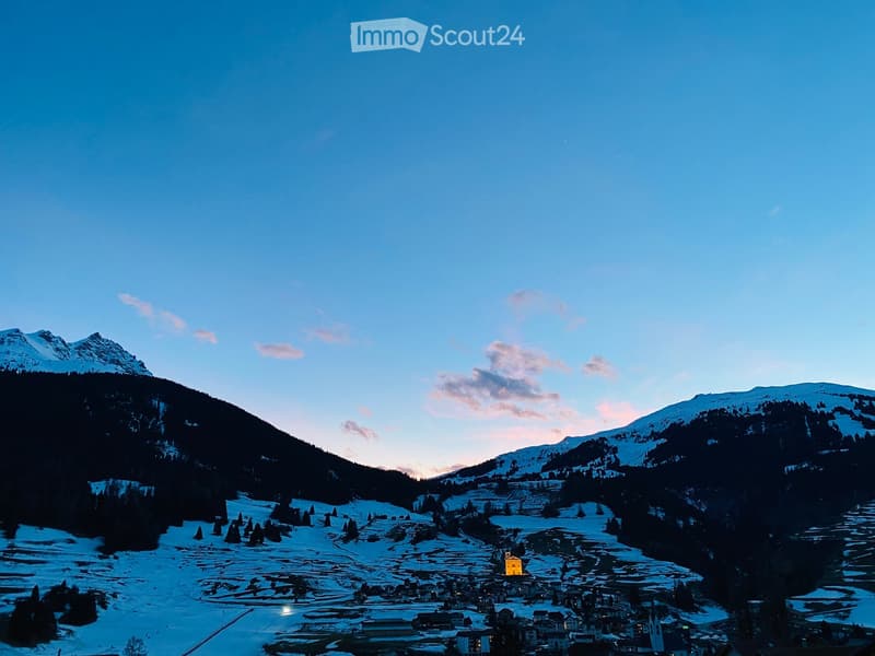 A picturesque village nestled in a snow-covered valley with surrounding mountains, featuring scattered houses and a church illuminated against a twilight sky.