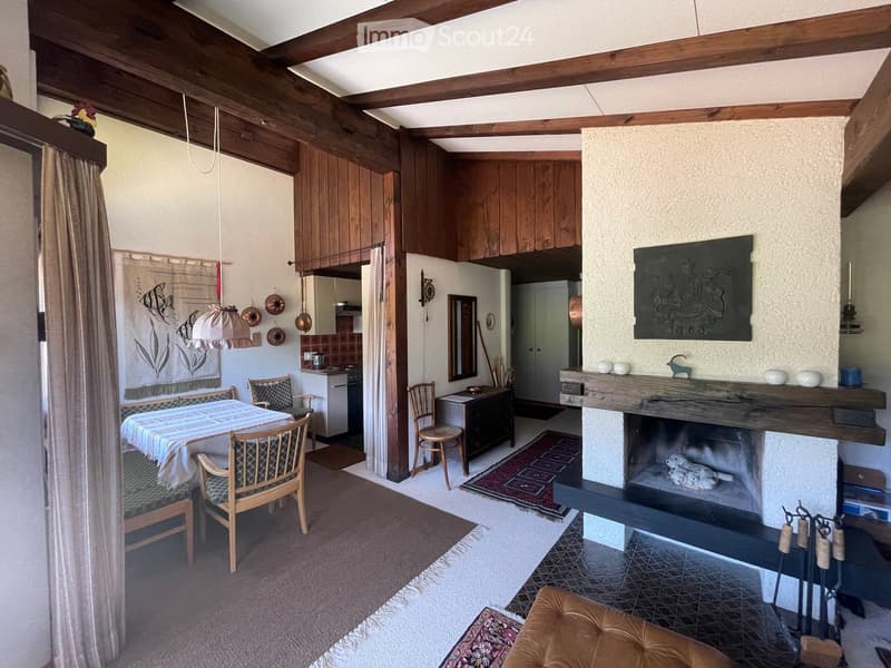 Cozy living and dining area with exposed wooden beams, a fireplace, a dining table with chairs, and a small kitchen area.