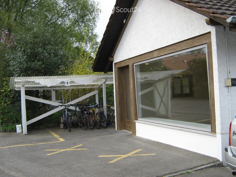A modern building with a covered parking area for bicycles and a car, featuring a large window and a white exterior.
