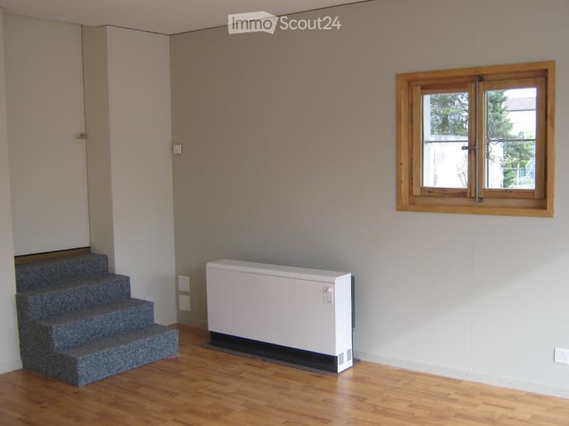 Empty room with wooden flooring, grey carpeted stairs, white radiator, and a window with wooden frame and metal security bars.