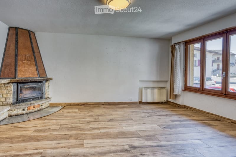 Empty room with wooden flooring, a stone fireplace with a metal hood, large windows with wooden frames, and a radiator on the wall.