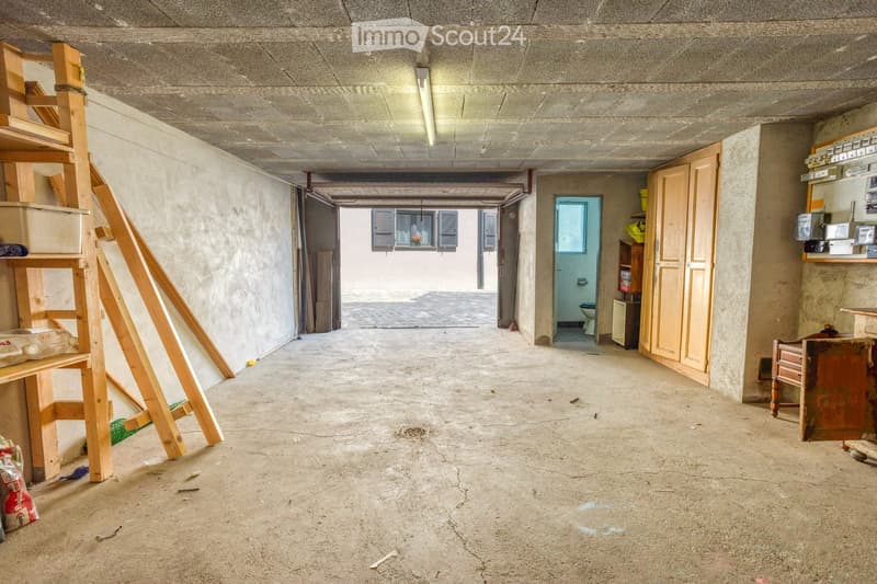 Empty room with bare concrete walls and floor, wooden ladder, and a partially visible bathroom area in the background.