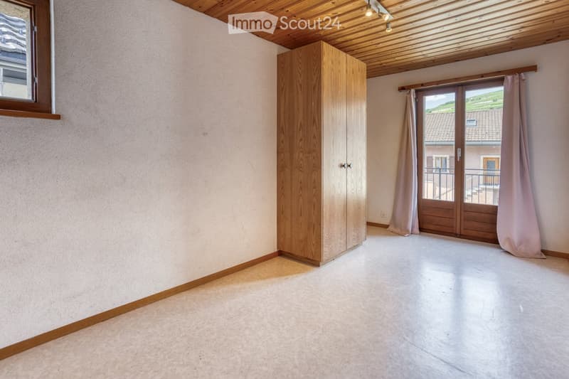 Empty room with light-colored walls, wooden floor, and a large wooden wardrobe. A sliding glass door with curtains leads to a balcony or outdoor area.