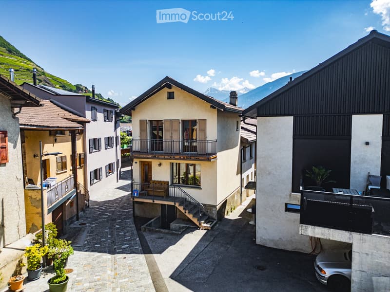 A charming European village street with traditional houses, cobblestone pavement, and a scenic mountain backdrop.