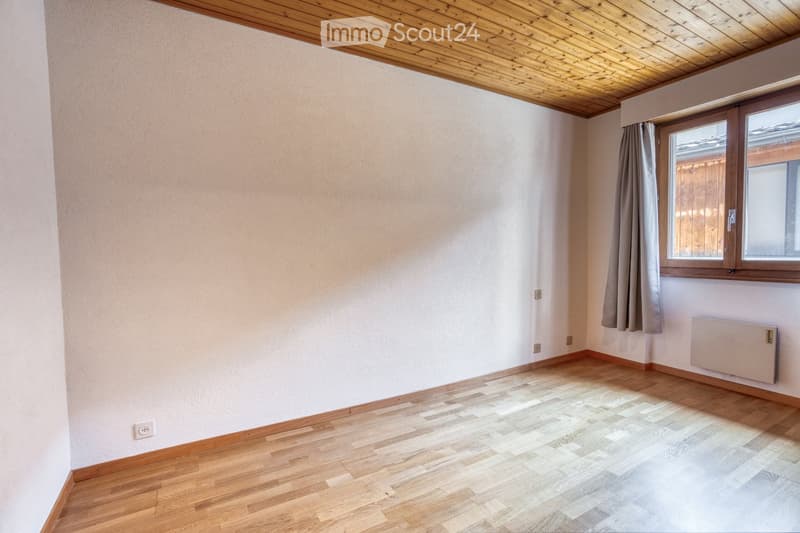 Empty room with wooden flooring, white walls, and a window with curtains. The room features a wooden ceiling and a radiator on one wall.