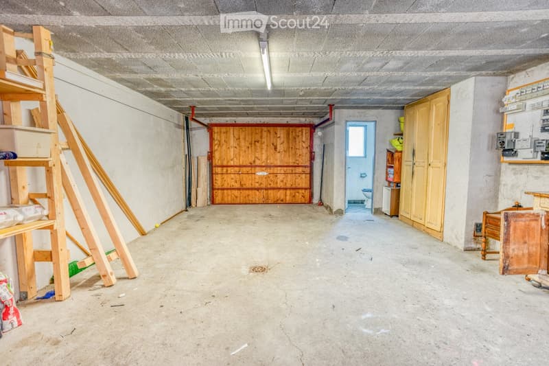 Empty garage with concrete floor, wooden door, and a small adjacent bathroom. Shelves and tools are visible on the left side.