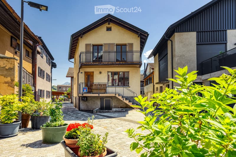 A charming two-story building with a balcony and a staircase leading to the entrance, surrounded by potted plants and greenery in a cobblestone courtyard.