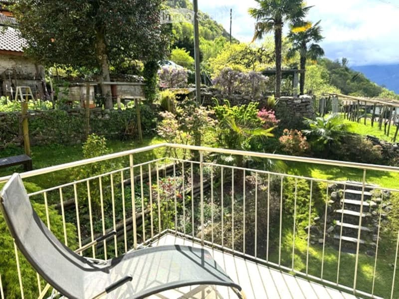 Balcony with a metal railing, a lounge chair, and a small table overlooking a lush, green garden with trees and a stone pathway.