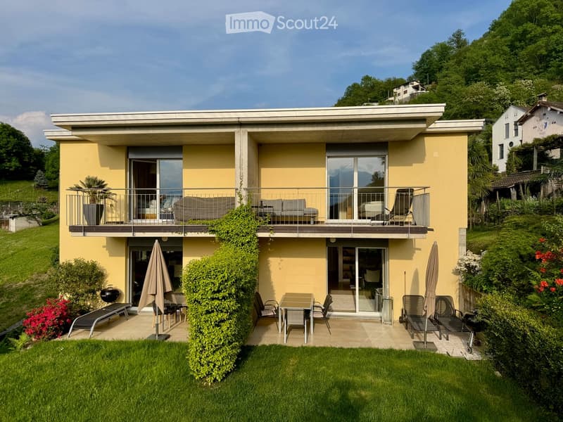 Two-story modern house with yellow facade, large windows, and balconies. Features include a well-maintained garden, patio seating, and outdoor dining area.