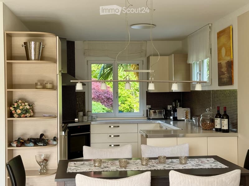 Modern kitchen with white cabinets, stainless steel appliances, and a dining area with a table and chairs. Shelves display decorative items, including ducks, flowers, and glassware.