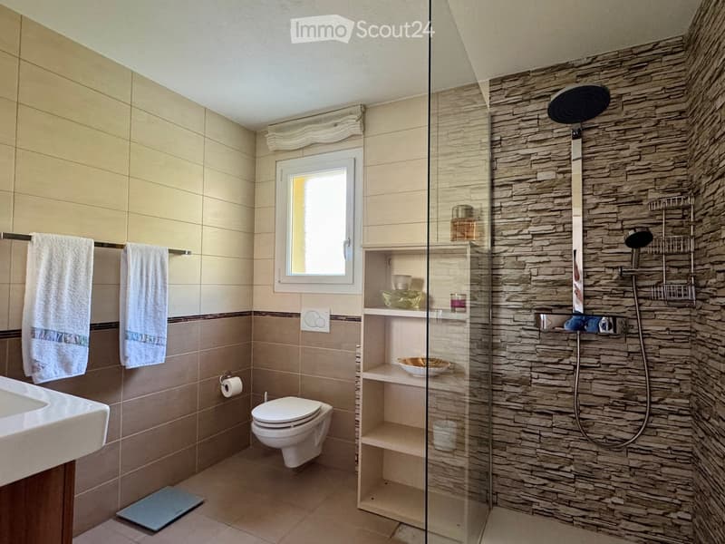 Modern bathroom with beige tiles, a glass-enclosed shower with stone walls, a toilet, a sink, and a window with a towel rack.