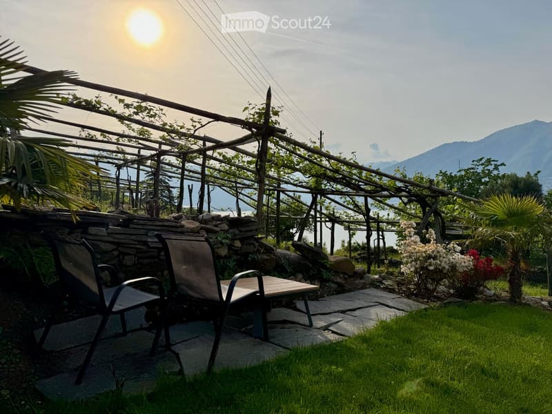 Garden with a stone wall, wooden pergola, two chairs, a small table, and lush greenery, overlooking a scenic mountain and lake view.