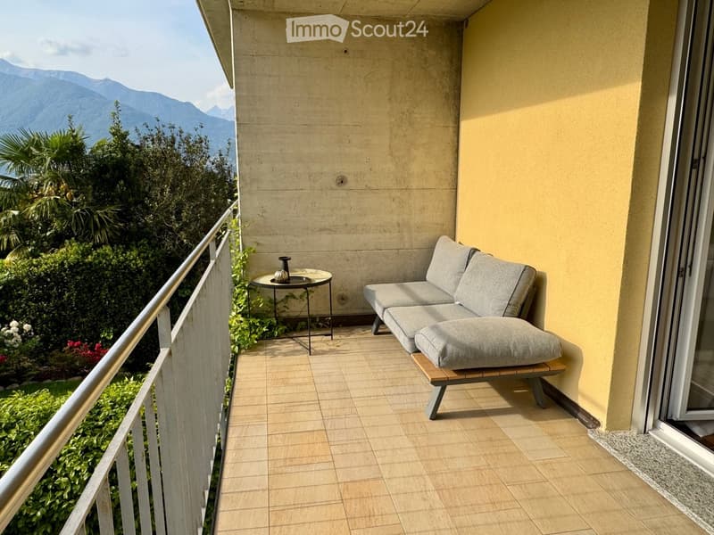 Balcony with a view of mountains, featuring a grey sofa, a small round table, and a decorative vase.